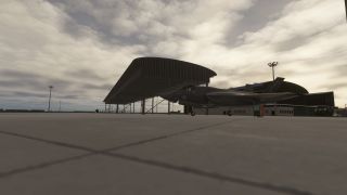 A gray fighter jet sits with landing gear extended on a concrete apron beside an open-sided shelter under cloudy skies.