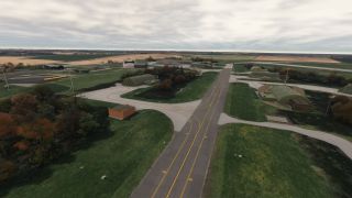 A yellow-marked taxiway runs between earth-covered aircraft shelters, with grassy areas, autumn trees, and surrounding farmland beneath an overcast sky.