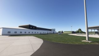A long white airport building borders a grassy area, with concrete pavement, green arched shelters, and tall light poles nearby.
