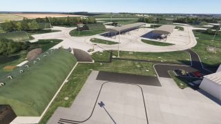 An airfield features green arched aircraft shelters, open-sided canopies, curving taxiways with yellow markings, and parked vehicles beside grassy areas.