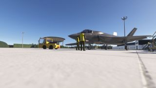 A gray fighter jet sits on the apron beside two ground crew in yellow vests and a yellow tow tractor.