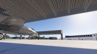 Two gray fighter jets sit beneath open-sided shelters, with ground crew beside the nearer aircraft and airport buildings across the apron.