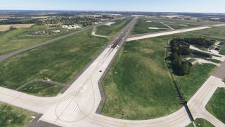 A runway with white threshold markings and dark tire streaks extends between grassy infields, intersecting taxiways, and arched aircraft shelters.