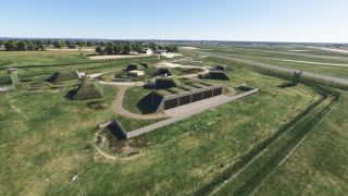 Grass-covered bunkers occupy a fenced airfield compound with watchtowers, access roads, and runways stretching across the background.