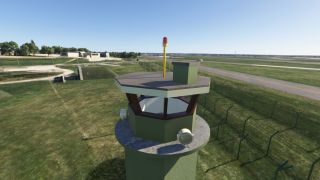 A green airport observation tower with spotlights and a red rooftop beacon stands beside perimeter fencing and a taxiway.
