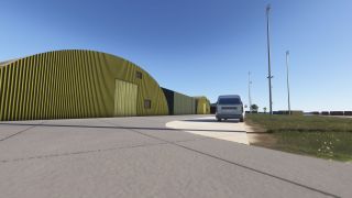 A silver vehicle sits beside green corrugated hangars with curved roofs, while tall floodlight poles line the grassy verge.