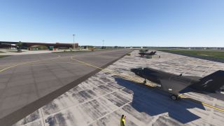 Three dark fighter jets sit on a concrete apron with yellow markings, ground equipment, and hangars beneath a clear blue sky.