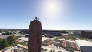 A tall brick tower topped with antennas overlooks airport hangars, paved service areas, and scattered trees under a clear blue sky.