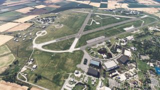 An aerial view shows an airfield with intersecting runways, curved taxiways, hangars, and surrounding patchwork farmland.