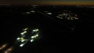 An airport at night shows illuminated runway edges, green taxiway lights, floodlit buildings, and distant city lights beneath a dark orange horizon.