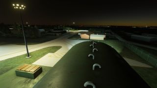 Floodlights illuminate airport taxiways beside an arched shelter with curved roof vents, low buildings, and a perimeter fence at night.