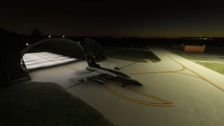 A military jet with underwing tanks sits outside an illuminated arched shelter at night, amid curved yellow taxiway markings.