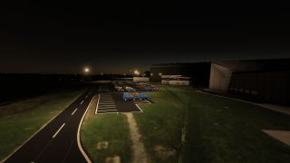 An illuminated parking lot contains cars and blue vans beside large hangars, a perimeter fence, and an access road at night.