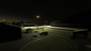 Floodlights illuminate an airport service area at night, with parked trucks and cars beside large buildings and an open-sided shelter.