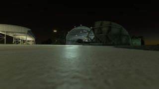 A military jet sits inside an illuminated arched shelter at night, beside an open-sided hangar and a curved yellow taxiway line.