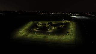A floodlit, fenced compound contains bunker-like structures beside green taxiway lights and a lit runway at night.