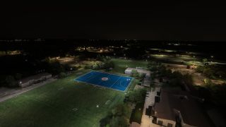 An aerial nighttime view shows a blue sports court with a central roundel, grassy playing fields, surrounding buildings, and illuminated roads.