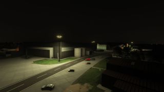 Floodlights illuminate airport hangars, cylindrical storage tanks, and parked cars beside a marked service road at night.