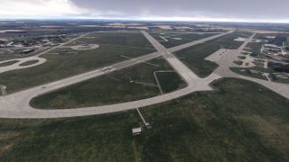 Intersecting runways and curved taxiways span a grassy airport, with parked aircraft, hangars, and surrounding farmland beneath cloudy skies.