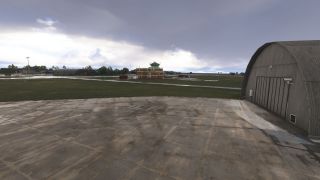 A curved-roof hangar borders a weathered concrete apron, with a brick control tower across the grass beneath gray clouds.