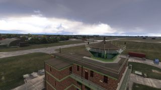 A brick control tower with green trim overlooks taxiways, grass fields, and arched hangars beneath heavy gray clouds.