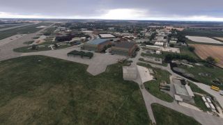 An aerial view shows airport hangars, parked aircraft, taxiways, and service buildings surrounded by farmland beneath an overcast sky.