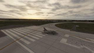 A gray fighter jet sits on runway threshold markings beside curved taxiways, with low hangars and a cloudy sky beyond.