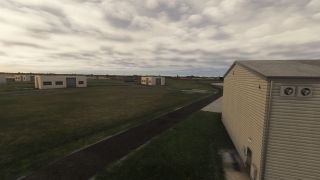 Low airport buildings stand across grassy grounds beside a narrow paved lane, with a large beige hangar beneath overcast skies.