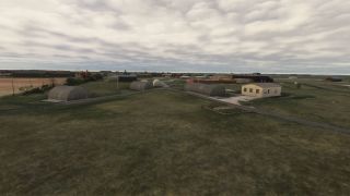 Several arched hangars and a rectangular building stand beside paved access roads across a grassy airfield under overcast skies.