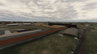 An airfield contains arched hangars, rectangular buildings, and paved access roads amid grassy areas beneath extensive cloud cover.