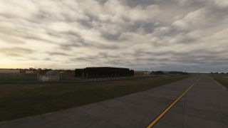 A concrete taxiway with a yellow centerline runs beside fenced airport buildings and grassy verges beneath extensive cloud cover.