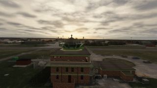 A brick airport control tower with green-framed windows overlooks taxiways, hangars, and parked cars beneath a cloudy sky.