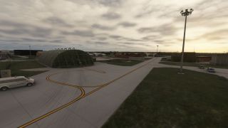 Curving yellow taxiway lines lead past an arched aircraft shelter, with service vehicles and tall floodlight poles beneath a cloudy sky.