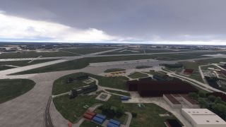 Intersecting runways and taxiways extend across an airport, with hangars, service buildings, and yellow ground equipment beneath dark clouds.