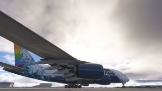A four-engine airliner with blue geometric livery and a rainbow tail stands on the apron, viewed from beneath its wing.