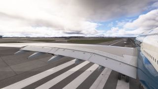 A double-deck airliner sits over runway threshold markings with wing flaps extended, landing gear visible, and airport buildings beneath cloudy skies.