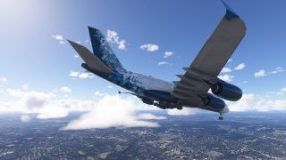 A four-engine airliner with blue geometric livery flies above urban terrain and scattered clouds with its landing gear extended.