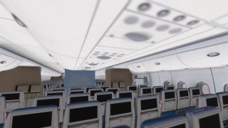 An empty passenger cabin has rows of seats with black seatback screens, closed overhead bins, and oval windows along the right wall.