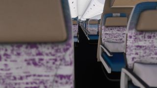 An empty aircraft cabin has rows of blue and purple patterned seats, beige headrests, and overhead bins above a narrow aisle.