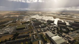 An aerial view shows airport hangars, parked aircraft, and reflective aprons beside runways, with surrounding fields beneath low clouds.