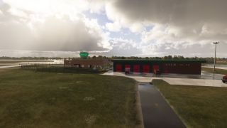 An airport fire station has red garage doors and parked fire engines beside a green-topped control tower and wet pavement.