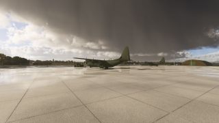 Two green military transport aircraft sit on a wet apron beneath dark clouds, with ground equipment and a curved-roof hangar nearby.