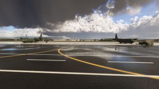 Two green four-engine transport aircraft sit on a wet apron beneath dark clouds, with a tow tractor beside the right aircraft.