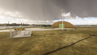 An arched hangar with green doors stands beside a floodlight mast, fenced equipment, and a yellow airport tug under dark clouds.