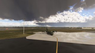 An olive-green four-engine turboprop transport sits on a wet apron beside a curved yellow taxi line beneath dark clouds.