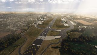Runway 07 extends between curved taxiways, hangars, and parked aircraft, with residential streets to the left and farmland beyond.