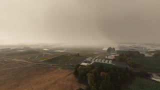 An airport runway fades into thick haze, with yellow taxiway markings, hangars, perimeter roads, and trees visible in the foreground.