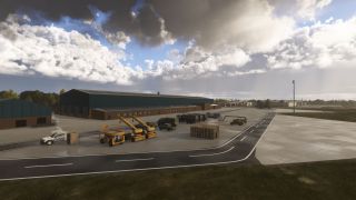Yellow lifting equipment, tanker trucks, and stacked pallets occupy an airport service yard beside large green hangars under broken clouds.