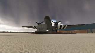A twin-engine Dakota with black-and-white wing stripes stands behind an Air Despatch memorial stone beside a hangar under dark clouds.