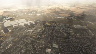 An airport lies between dense residential streets and farmland, with a long runway, hangars, and several aircraft parked on broad aprons.